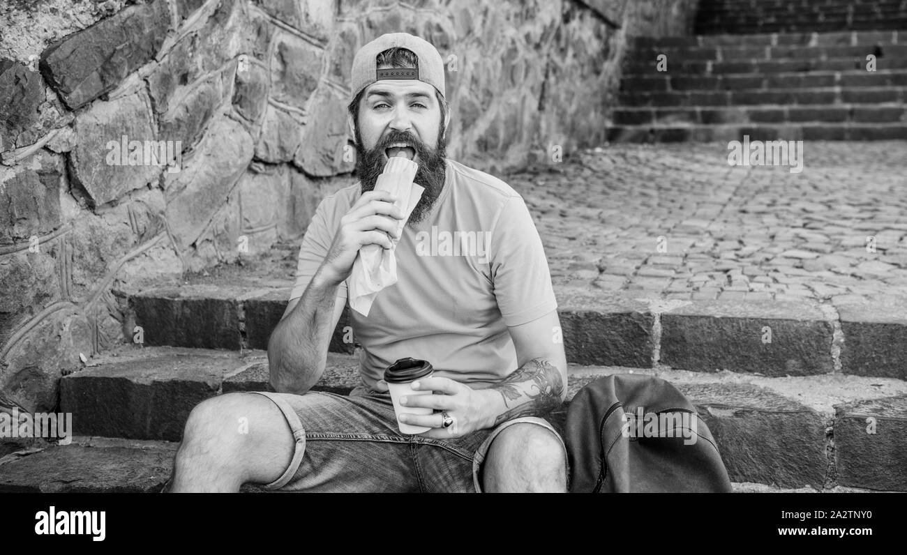 Uomo Barbuto godetevi uno spuntino veloce e bere bicchiere di carta. Cucina di strada così buona. Uno stile di vita urbano nutrizione. Carefree hipster mangiare cibo spazzatura mentre sedersi sulle scale. Uomo affamato snack. Il cibo spazzatura. Guy mangiare hot dog. Foto Stock
