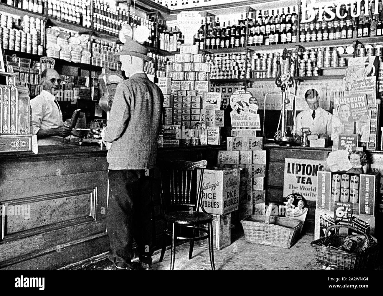 Negativo - all'interno di un negozio di alimentari, Ballarat, Victoria, circa 1930, un cliente e due assistenti shop all'interno di un negozio di alimentari. Prodotti come il 'Kellogg's bolle di riso " e " Lipton il tè" vengono pubblicizzati Foto Stock