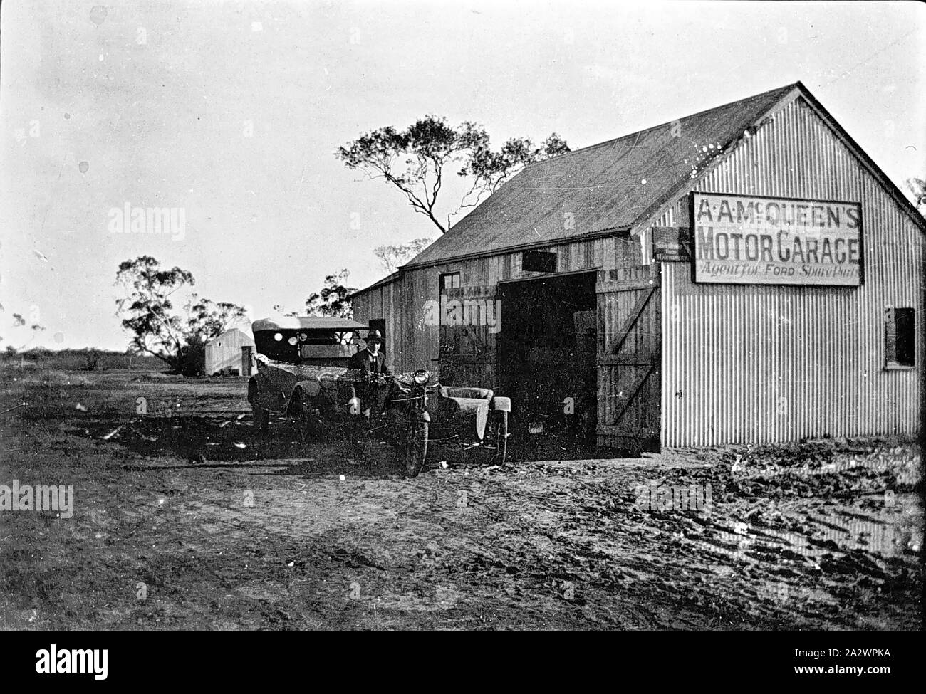 Negativo - R.A. McQueen il garage, Manangatang, Victoria, 1922, il motore il garage è un di legno e di ferro ondulato apparentemente edificio situato in aperta campagna. Un uomo con un auto e ciclo motore sorge nella parte anteriore dell'edificio Foto Stock