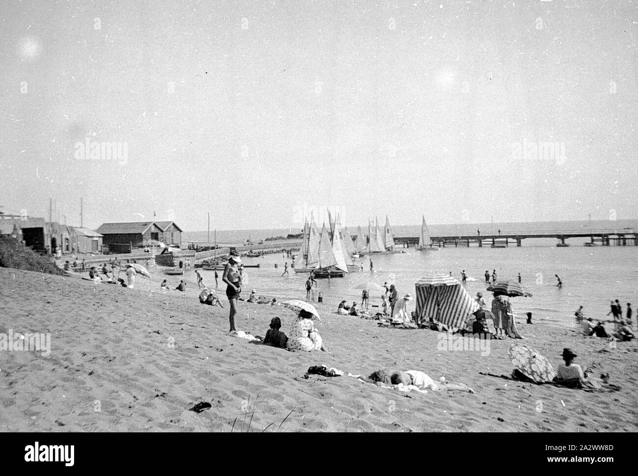 Negativo - Black Rock, Victoria, circa 1935, la spiaggia di roccia nera con barche a vela, i bagnanti e un molo. Ci sono persone sulla spiaggia con ombrelloni da spiaggia Foto Stock