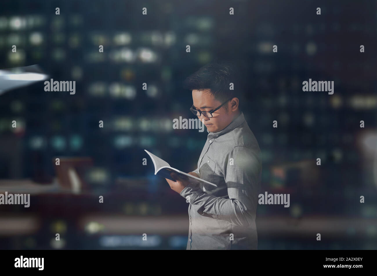 Una vista laterale colpo di un giovane uomo a maniche lunghe e bicchieri pacificamente la lettura di un libro all'interno di un ufficio scuro stanza a notte tempo. Foto Stock