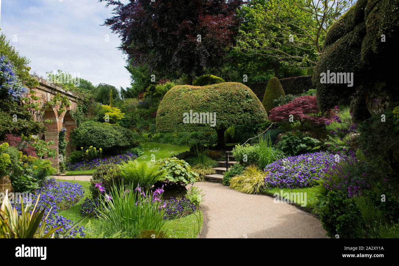 Il castello di Hever Castle Gardens, lussureggiante scenario e percorso curvo che conduce sotto un arco. Fiori. Blue sky. Formato orizzontale. N. persone. Foto Stock