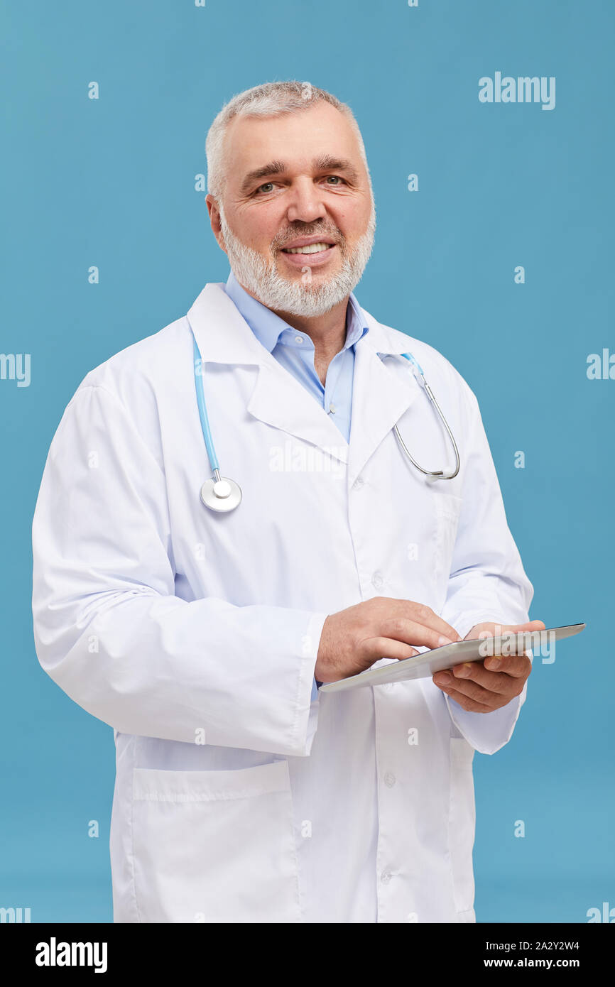 Ritratto di senior medico con i capelli grigi e in camice bianco sorridente alla videocamera mentre si lavora online su tablet pc su sfondo blu Foto Stock