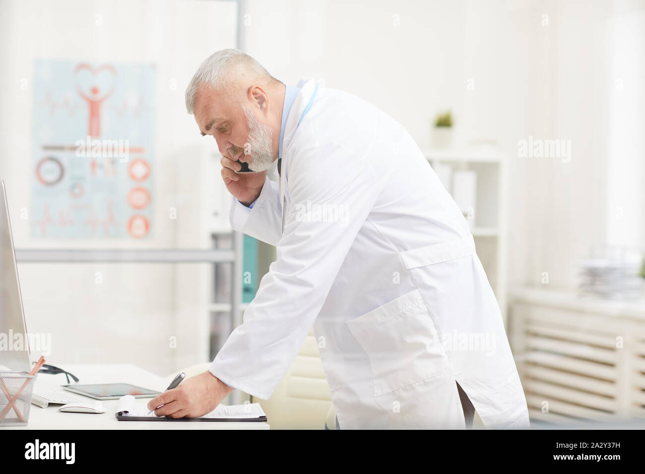 Senior medico in camice bianco concentrando sulla sua opera egli parlando al telefono cellulare e a rendere note in un documento di Office Foto Stock