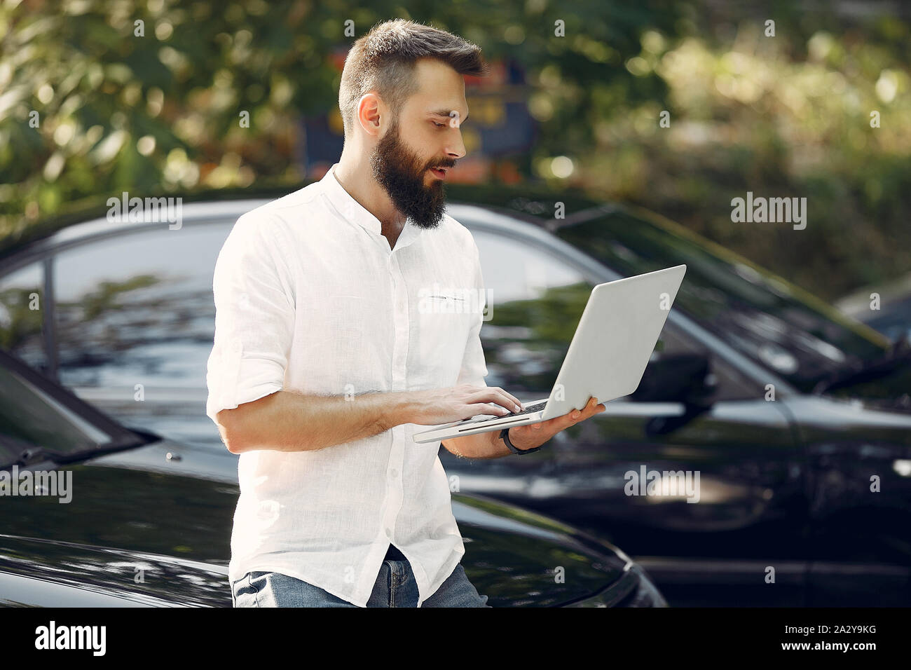 Uomo bello in una camicia bianca. Imprenditore lavora con il computer portatile. Uomo in piedi vicino a un auto Foto Stock