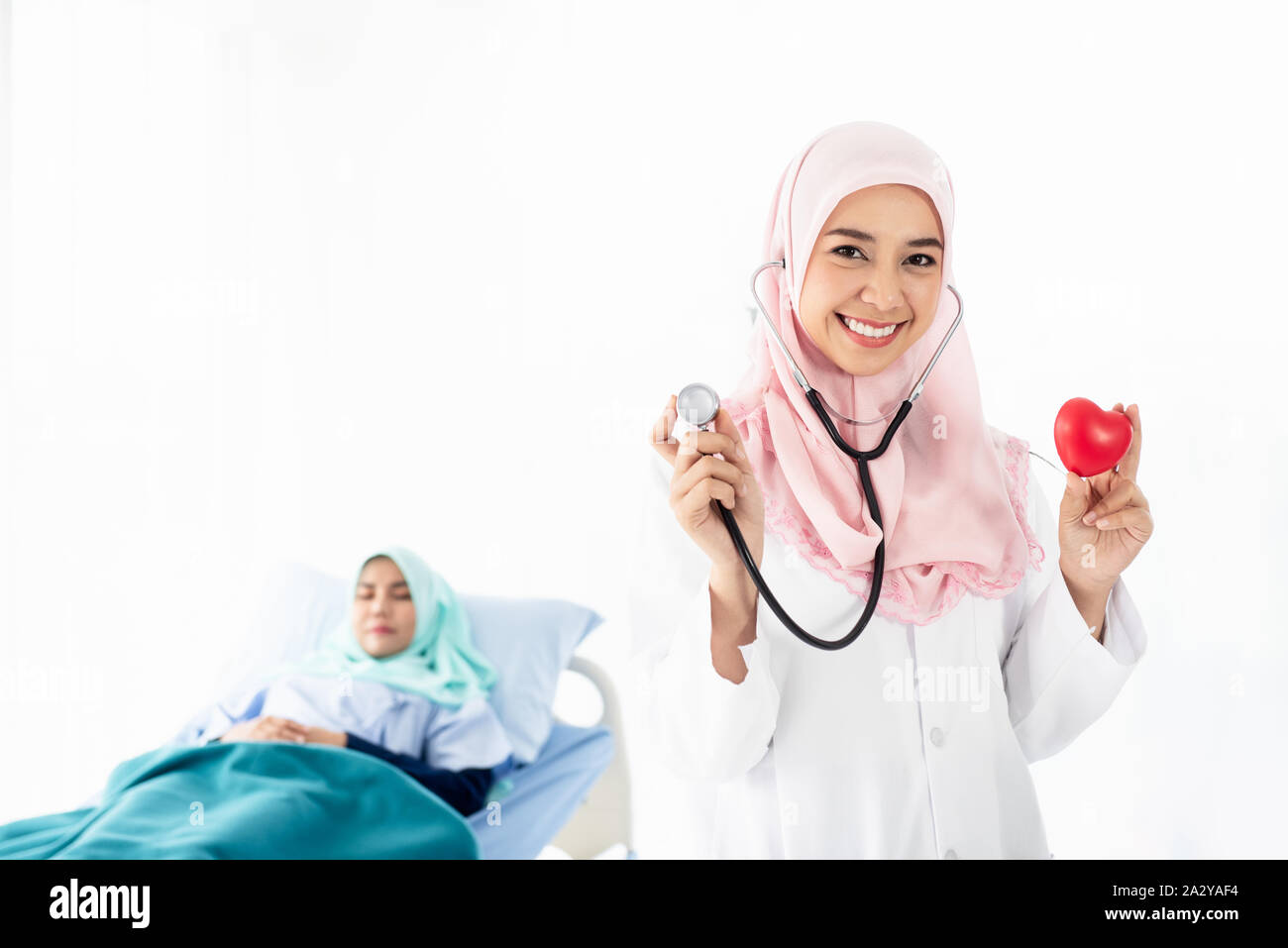 Ritratto di giovane e bella donna musulmana medico mostra un cuore rosso sul lato sinistro e indossa uno stetoscopio in piedi e sorrisi con pazienti giacente o Foto Stock