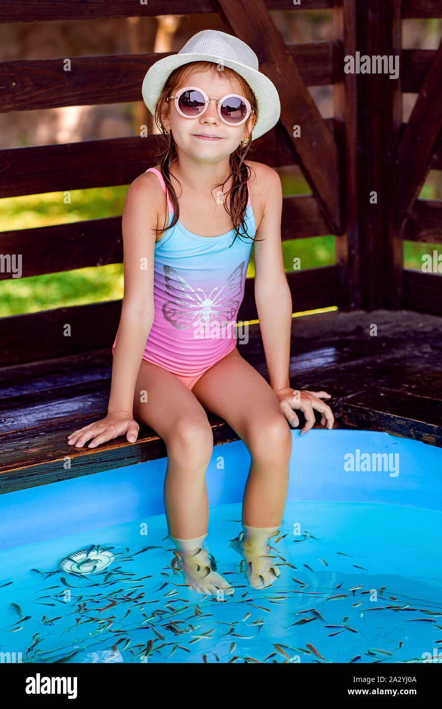 Ragazza giovane in cappello, costume da bagno che riceve massaggio con piccoli pesci. Peeling con pesce. Ragazza che gode di procedura medica. Massaggio plantare con pesce in un guardaroba dell'acquario. Procedura di Fish spa. Foto Stock