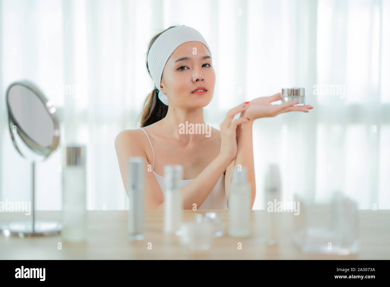 Ritratto di incantevole donna asiatica avente jar con crema per il viso in mano, utilizzando, applicando la lozione, godendo, toccando la sua guancia, con la cura della pelle sulla bottiglia ta Foto Stock