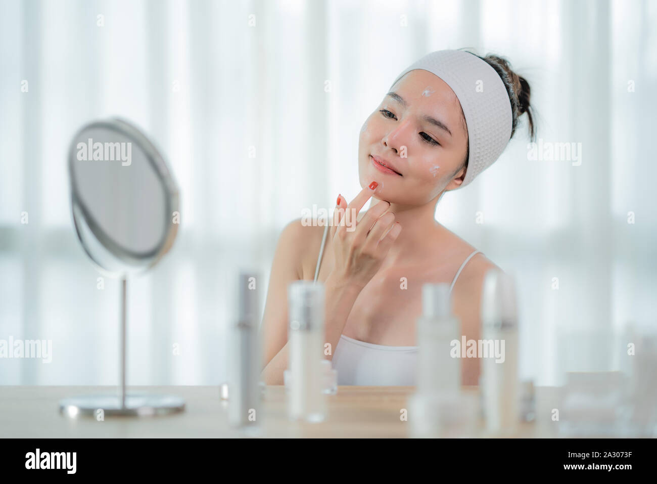 Ritratto di bella asiatica giovane donna prendersi cura della sua pelle seduto vicino a uno specchio in camera da letto con cura della pelle bottiglia sul tavolo, donna di toccare la faccia. Bellezza Foto Stock