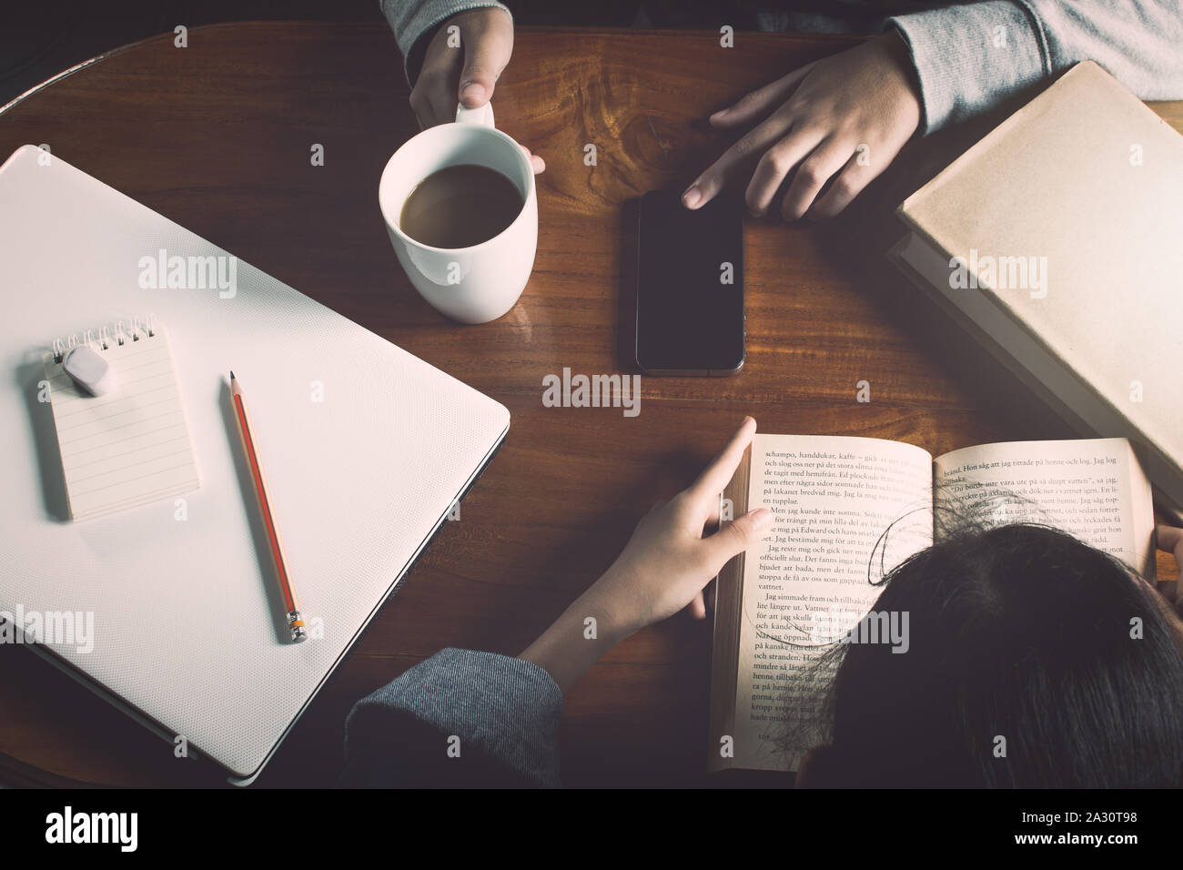 Ragazza la lettura di un libro e di un'altra ragazza utilizzando smart phone sul tavolo di legno con tazza di caffè Foto Stock