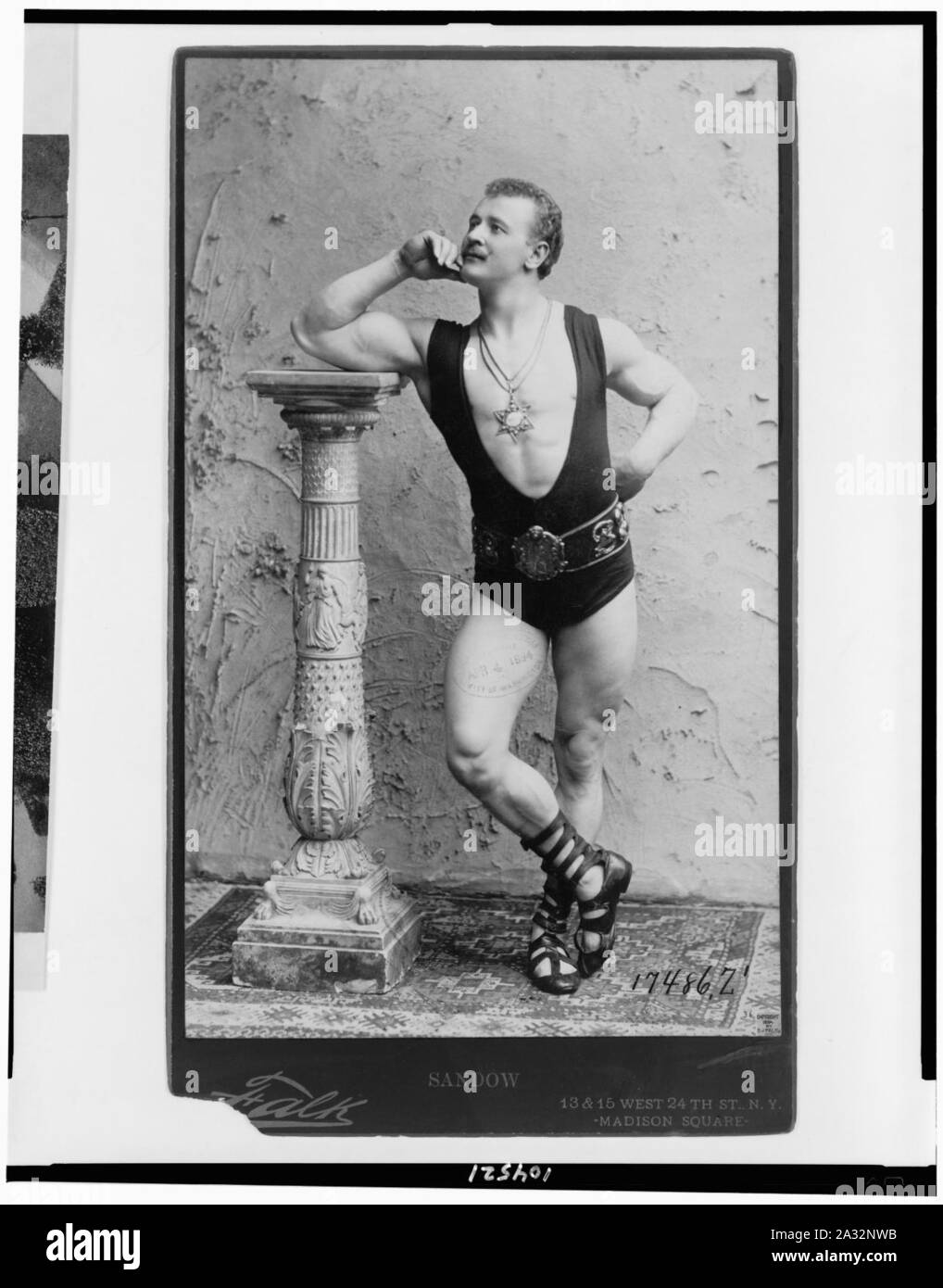 Eugene Sandow, ritratto a figura intera in piedi, poggiando su colonna, rivolto verso sinistra, indossando il wrestling leotard sandles romana, e la stella a sei punte, pendente Foto Stock