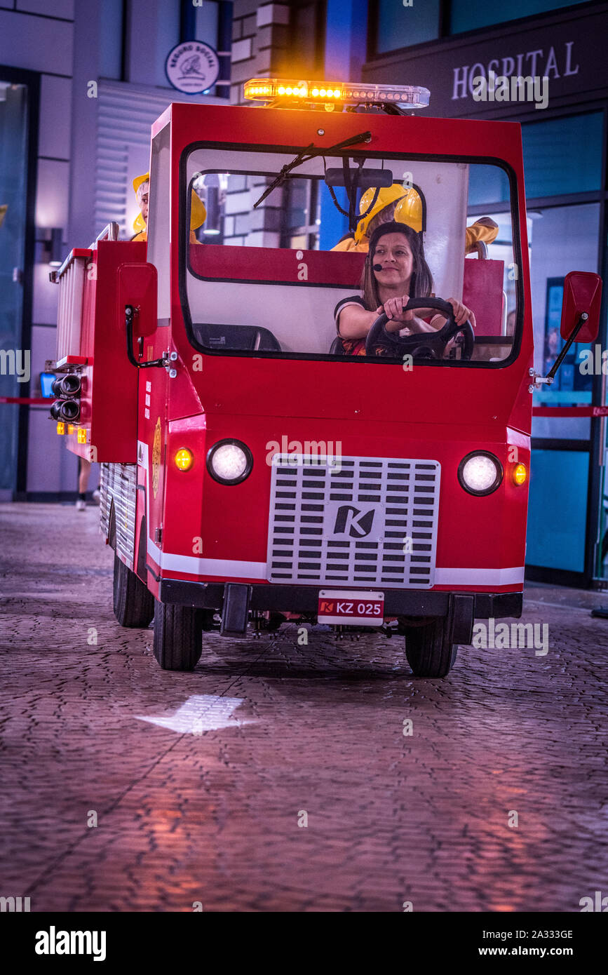 Vigile del fuoco rosso carrello con luci lampeggianti guidato da una donna sorridente sta prendendo una svolta a destra in una simulazione di città di Kidzania Foto Stock