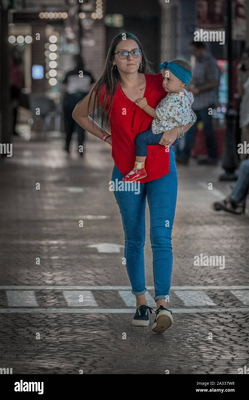 Una singola donna che indossa una maglietta rossa e blu jeans sta portando la sua figlia mentre si cammina su una strada pulita. Foto Stock