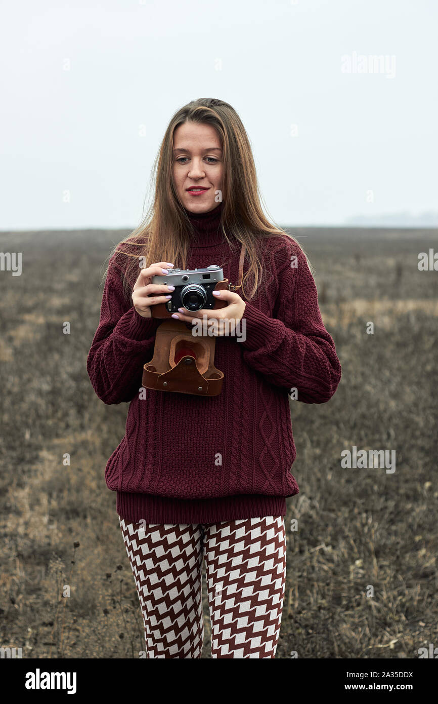 Giovane donna che indossa un maglione elegante e leggings con un pattern a mantenere vecchi film vintage fotocamera all'aperto nella pianura selvatici campo in autunno la nebbia fredda w Foto Stock