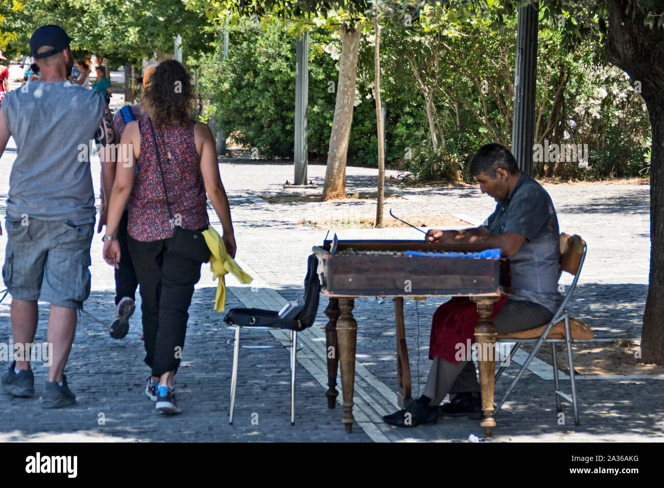 Atene, Grecia, giugno 04.2016.i turisti ad Atene a piedi passato un musicista di strada cercando di farli divertire da seduti all'ombra di un ulivo. Foto Stock