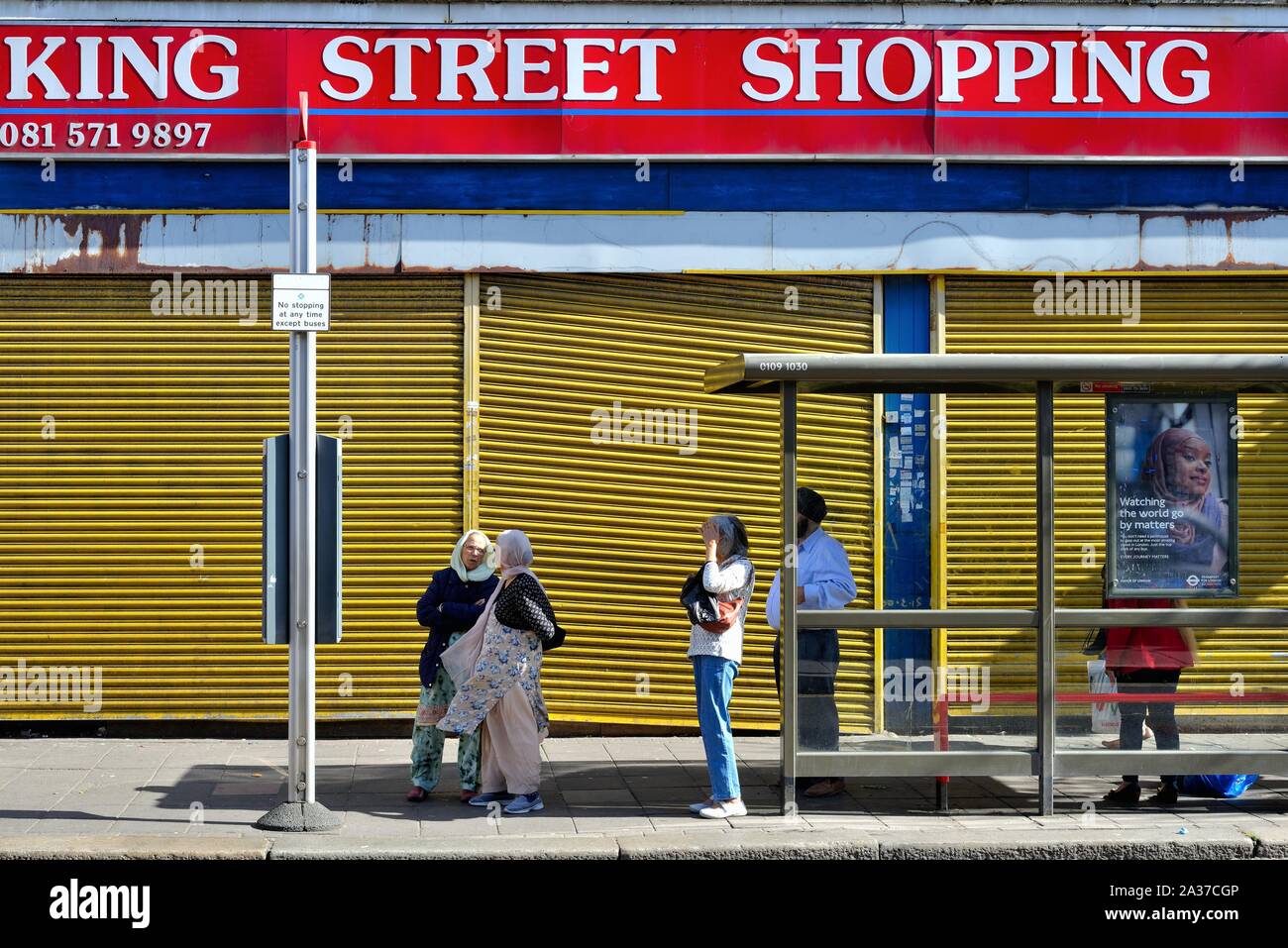 Razza mista persone in attesa alla fermata del bus sulla strada di Merrick, con una chiusa e parzializzato shop dietro, Southall West London Inghilterra England Regno Unito Foto Stock