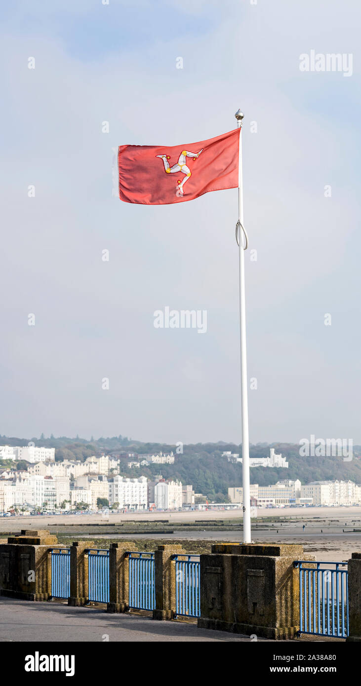 Isola di Man bandiera con triskelion contro uno sfondo rosso sollevato sul pennone sul lungomare di Douglas con Douglas, e la spiaggia in background. Foto Stock
