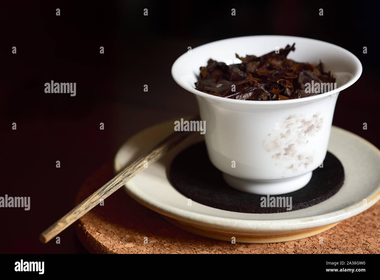 Primo piano di un tipico cinese teiera in porcellana bianca con infuso di tè nero foglie in esso su un piattino e un piatto di sughero con un vecchio bastone di legno contro Foto Stock