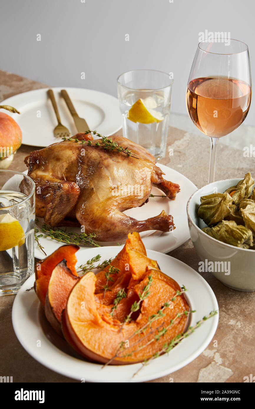 Grigliata di tacchino intero vicino al forno la zucca tagliata a fette e ciotola con physalis vicino a vetri con il vino rosato e acqua di limone isolato su grigio Foto Stock