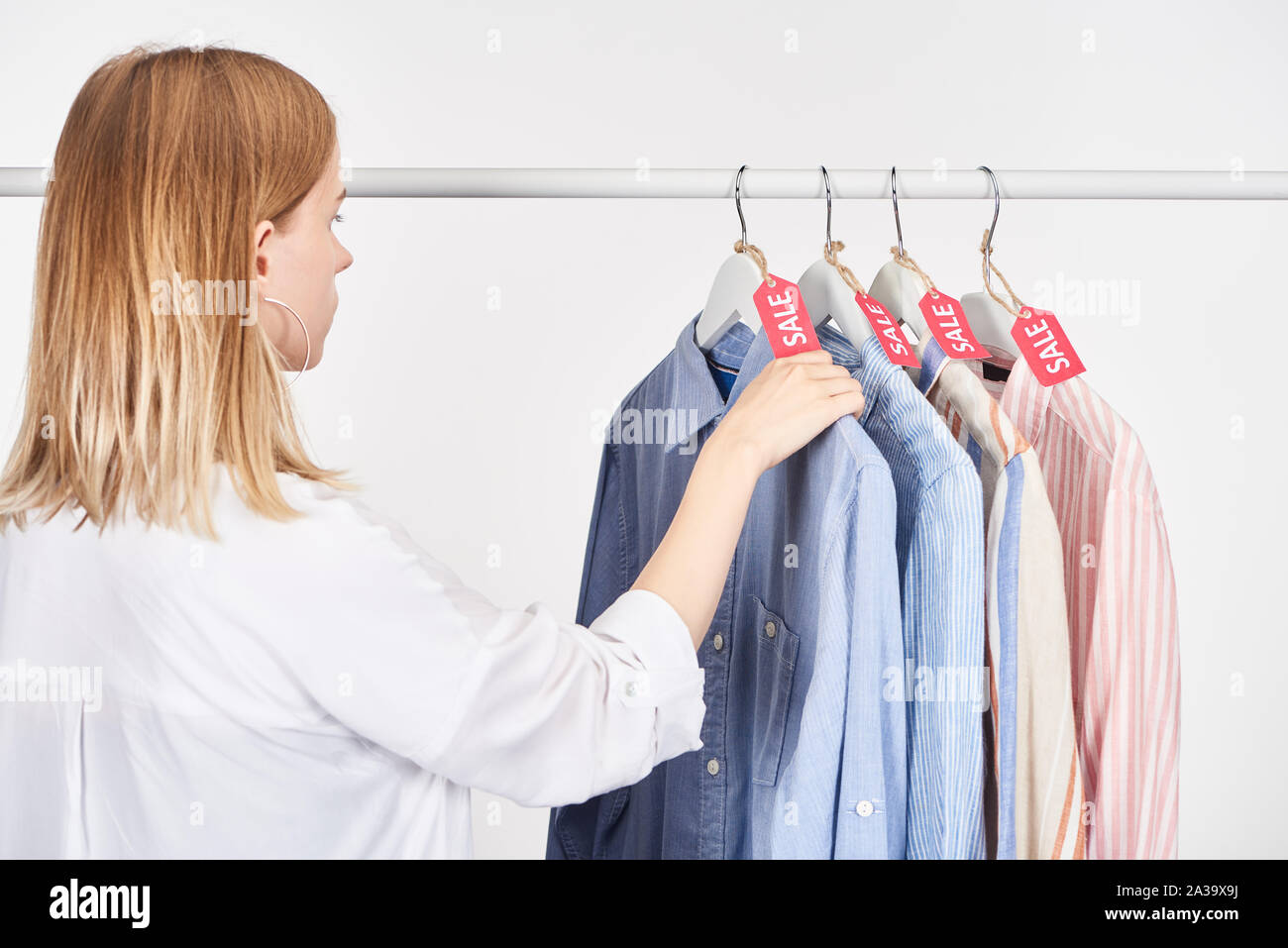 Donna bionda vicino eleganti camicie appeso con la vendita delle etichette isolato su bianco Foto Stock