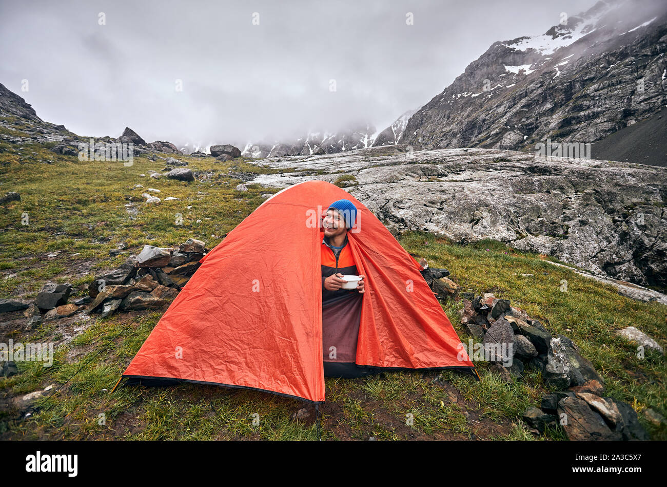 Felice turistico è di mangiare la sua colazione in tenda arancione nelle nebbiose montagne a piovosa mattinata a Karakol national park, il Kirghizistan Foto Stock