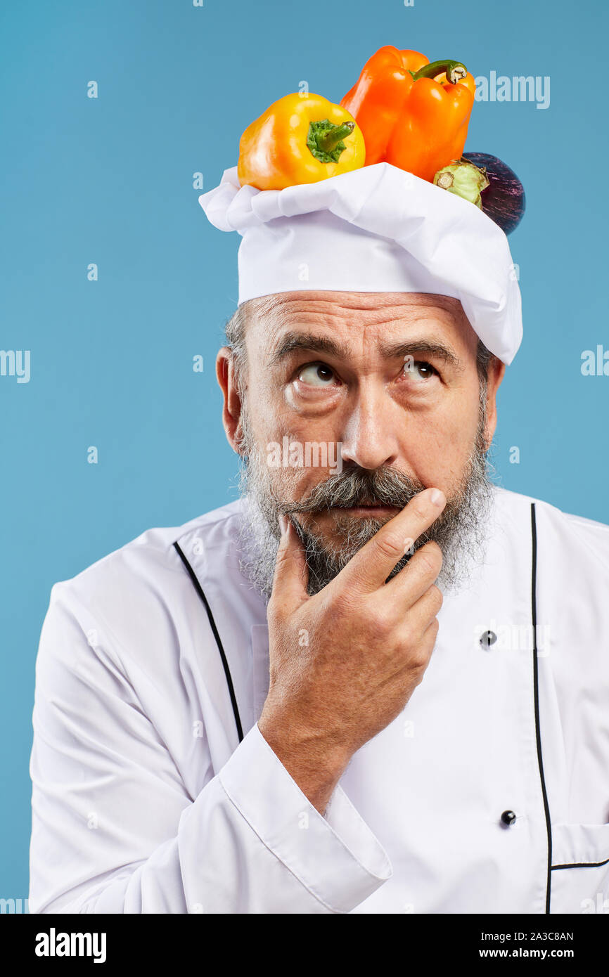 Ritratto del carismatico chef barbuto che guarda lontano pensively mentre posa contro sfondo blu con verdure fresche sulla parte superiore del cappello Foto Stock