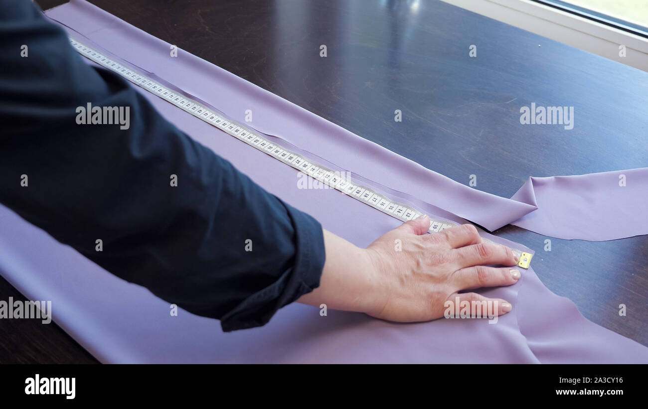 Taglio e cucito prodotti in sartoria aziendale. Sarta creando nuovi vestiti. Sarta rende un modello dal panno per cucire i vestiti, misure con nastro adesivo sul tavolo in officina, mani closeup. Foto Stock