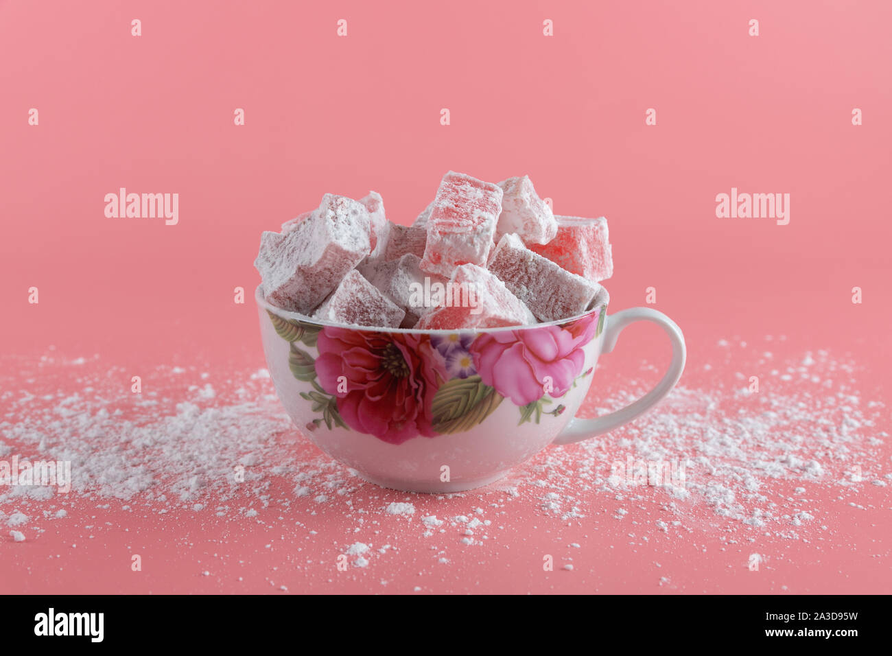Un boccale di bianco con fiori dipinti pieni di lokum, una delizia turca, su uno sfondo rosa e zucchero in polvere versato sul tavolo Foto Stock
