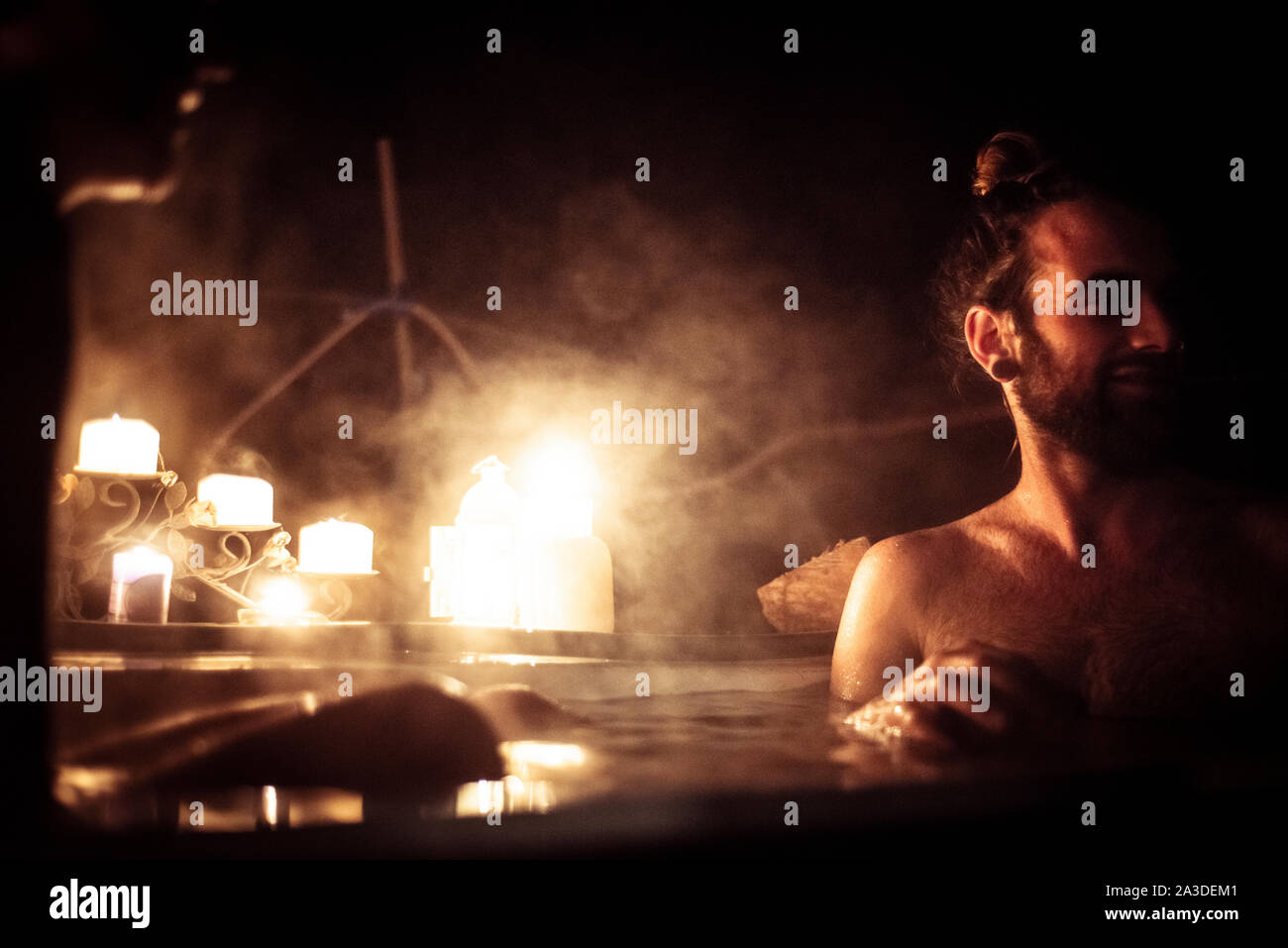 L'uomo con i capelli di bun siede in out door vasca da bagno al lume di candela Foto Stock