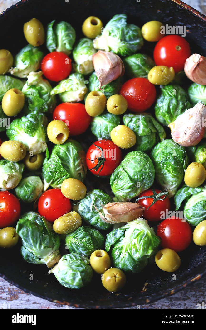 I cavoli di Bruxelles in un tegame con i pomodori ciliegia, aglio e olive prima della cottura. Cibo sano. Dieta vegana. Coppa vegetale. Foto Stock