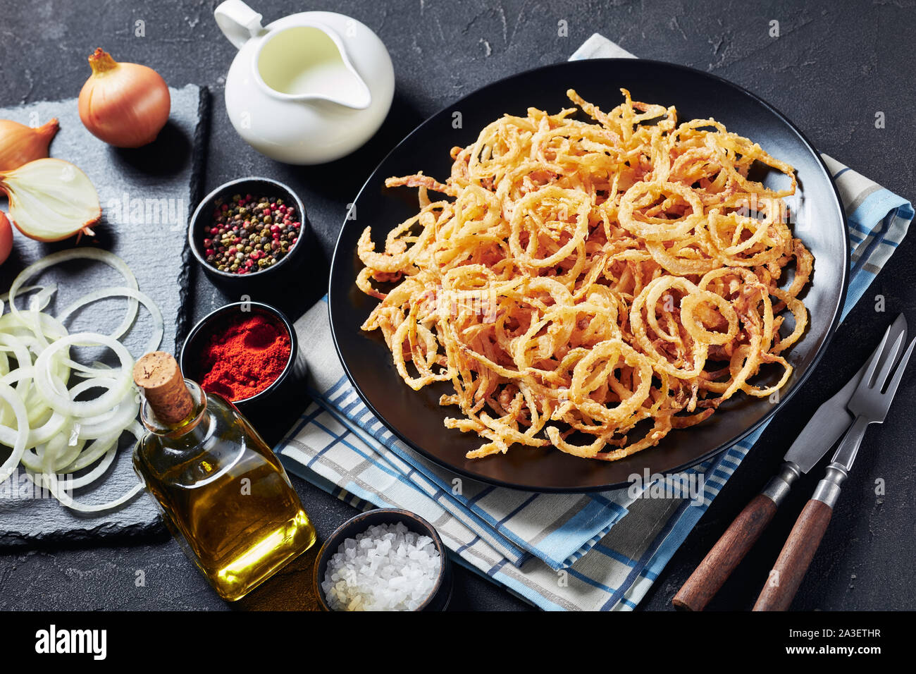 Close-up di croccanti anelli di cipolla fritti e stringhe con croccante di rivestimento su una piastra nera su una tabella di calcestruzzo con ingredienti, vista da sopra Foto Stock