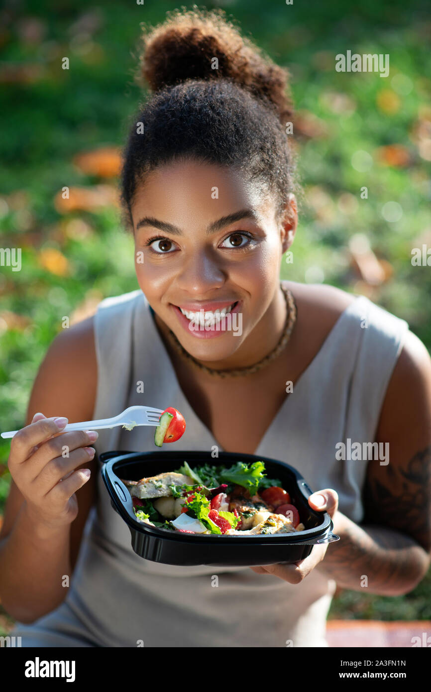 Dark-eyed donna sorridente mentre mangia squisita insalata di verdure Foto Stock