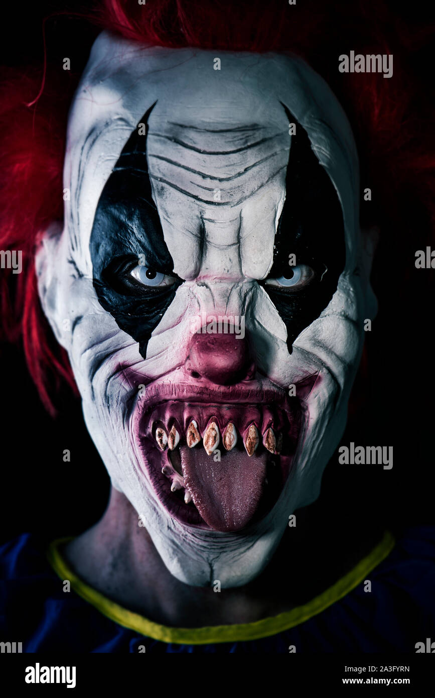 Primo piano di una paura male clown con i capelli rossi, bianco occhi, denti sanguinosi, attaccare fuori la sua lingua fissando l'osservatore con un aspetto minaccioso Foto Stock