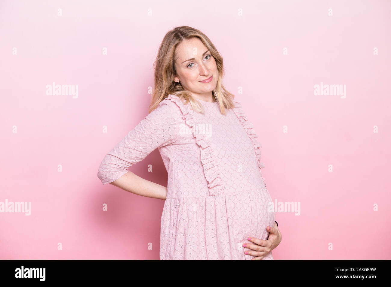 Giovane donna incinta indossando abiti rosa in piedi mentre tiene il suo ventre mantenendo una mano sulla sua schiena. Studio shot su sfondo rosa con spazio di copia Foto Stock