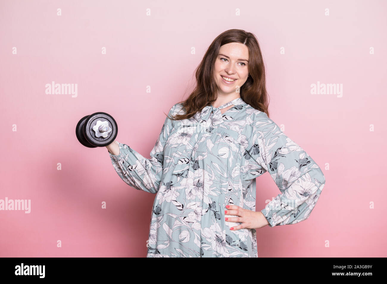 Felice giovane donna incinta indossa vestito blu fare esercizio di sollevamento pesi. Studio shot su sfondo rosa. Fitness e uno stile di vita sano durante il pr Foto Stock