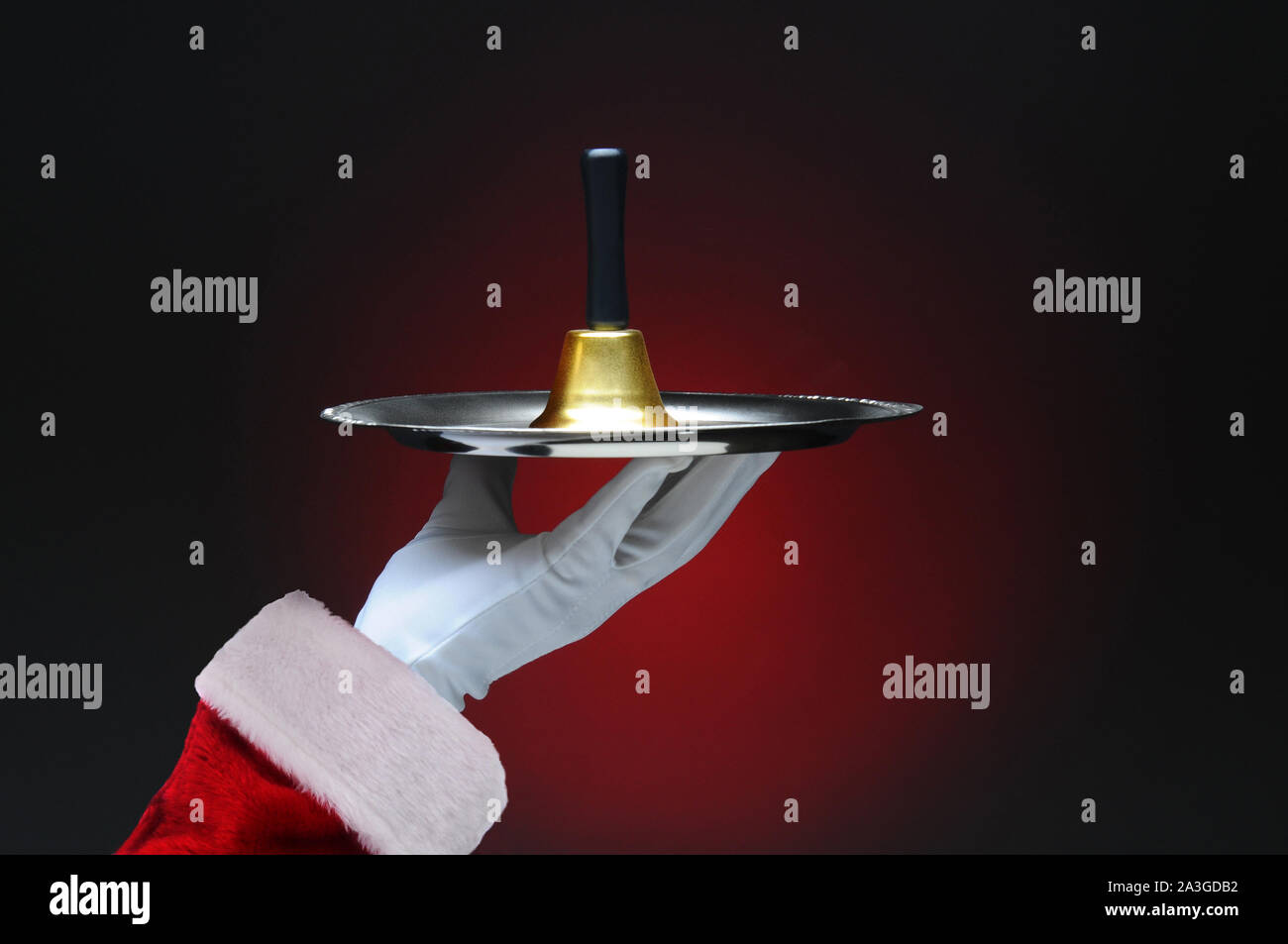 Primo piano di Santa Claus mano che regge una campana su un vassoio d'argento Formato orizzontale su una luce di colore rosso scuro dello sfondo. Foto Stock