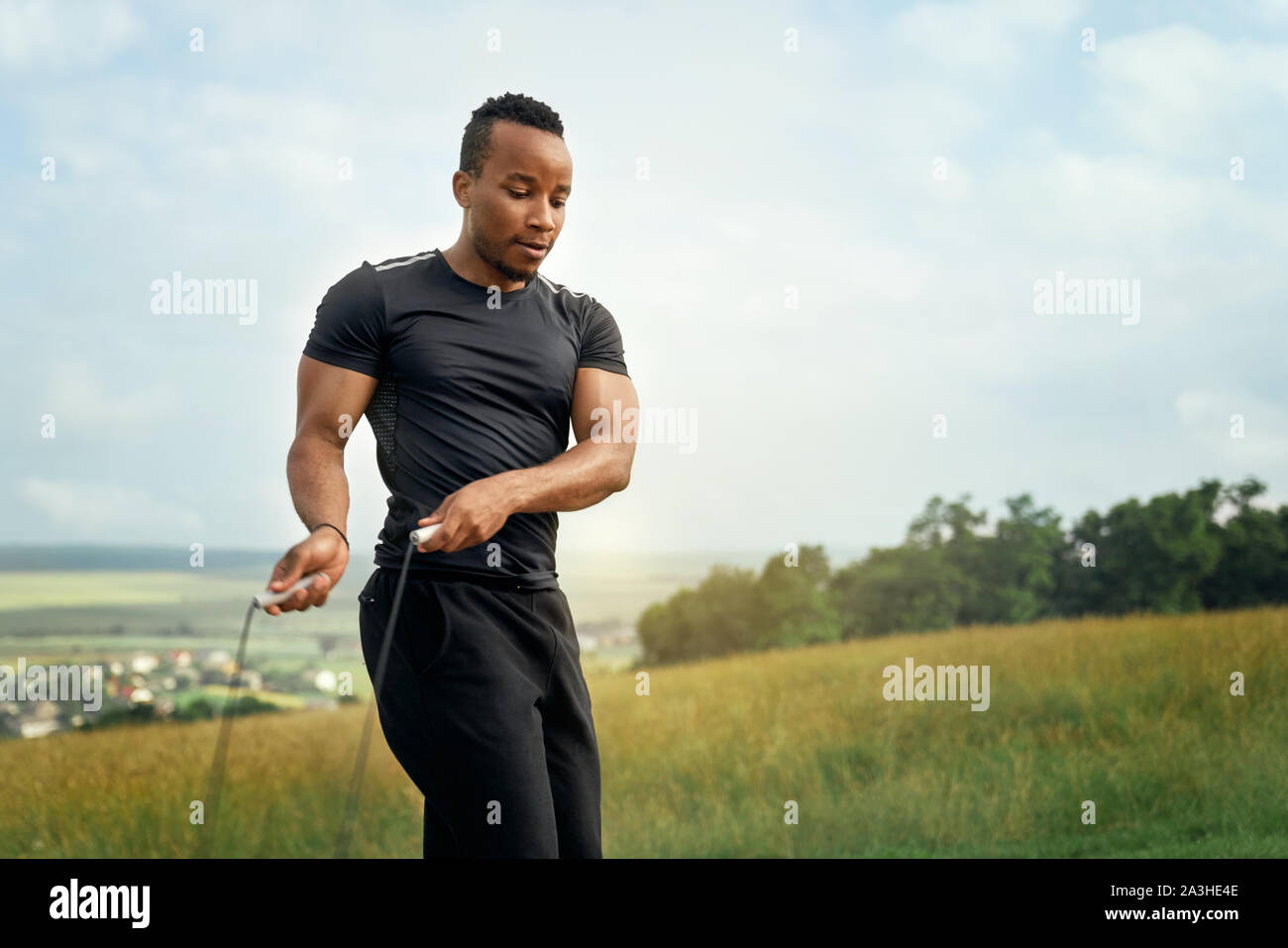 Muscolare, sportivo atletica formazione sulla natura. Bello atleta facendo esercizi con salto con la corda. Uomo sportivo ponendo il corpo sullo sfondo del cielo blu, guardando verso il basso. Foto Stock