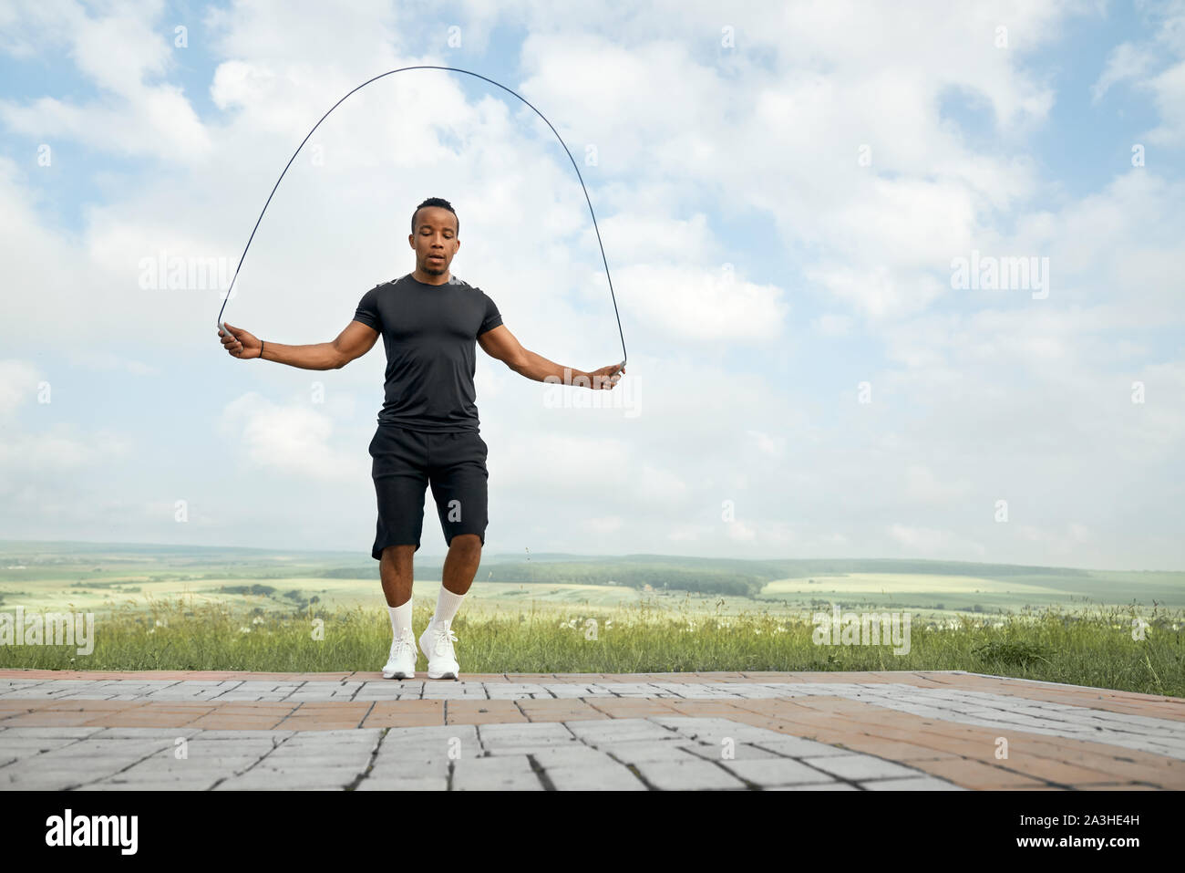 Attivo, bello, concentrata atleta jumping con salto con la corda all'aperto, guardando verso il basso. Uomo Forte con corpo sportivo in nero di t-shirt e shorts formazione, facendo esercizi. Foto Stock