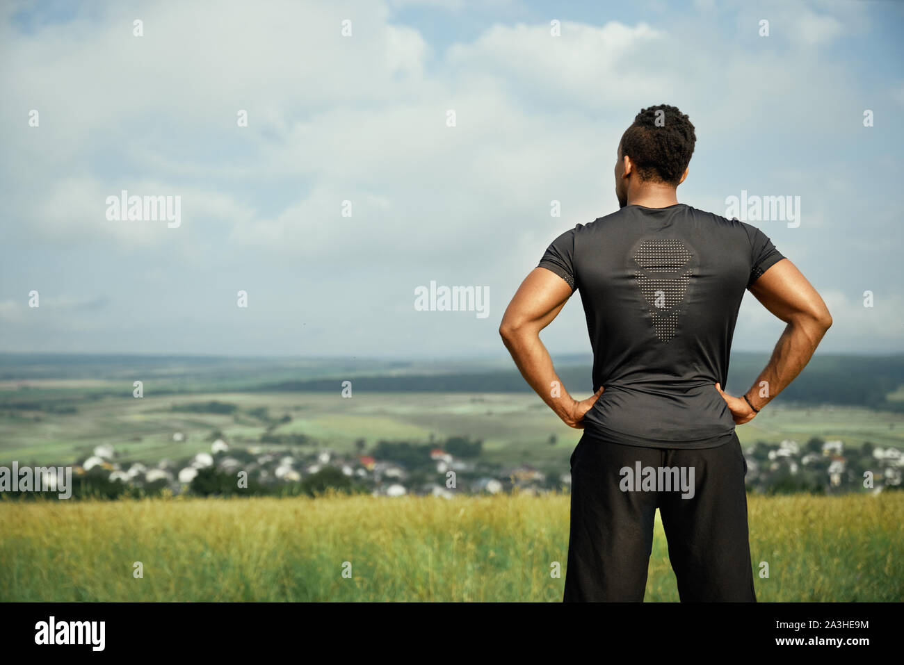 Vista posteriore del muscolare sportivo africani che indossa in nero di t-shirt e pantaloncini neri. Bello, forte, atletico uomo in piedi e in posa all'aperto. Atleta tenendo le mani sui fianchi. Foto Stock