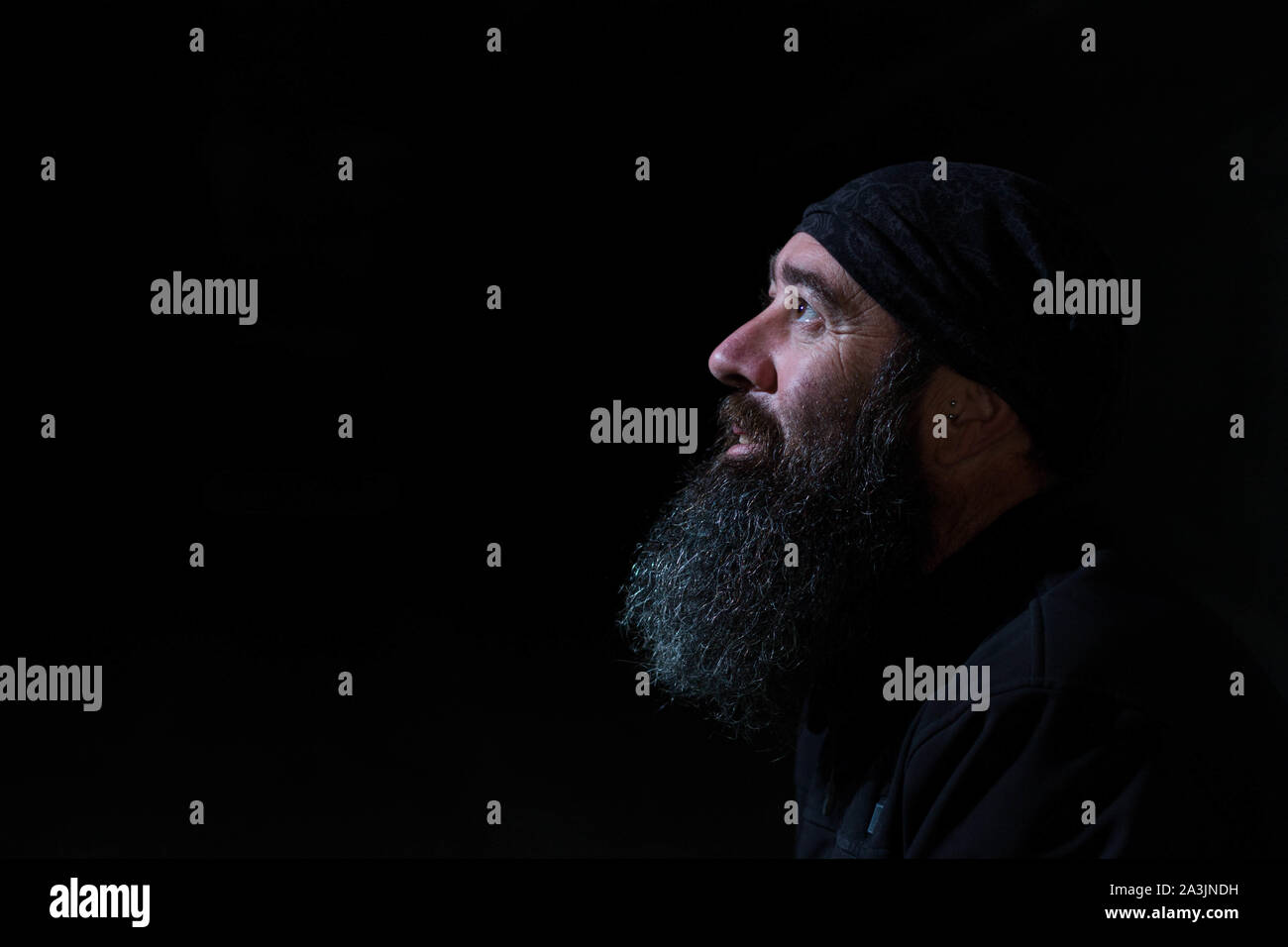 Studio shot di un lato verticale di un sorridente uomo barbuto con uno sfondo nero e guardando verso una luce con un foulard nero sulla sua testa e un earr Foto Stock