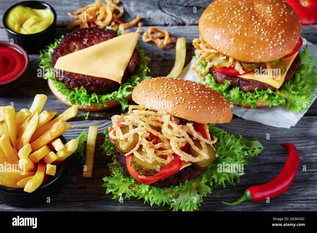 Tre cheeseburger con polpette di carne, formaggio cheddar, fritti e croccanti cipolle francese, lattuga, pomodori a fette, sottaceti su un vecchio tavolo di legno con musta Foto Stock