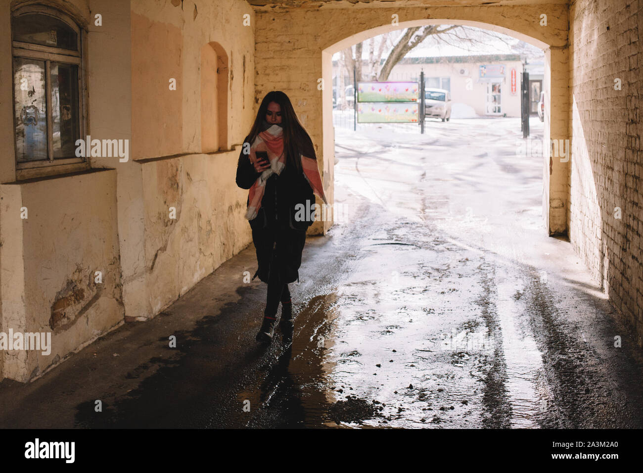 Giovane donna utilizzando smart phone mentre si cammina attraverso archway in città durante il periodo invernale Foto Stock