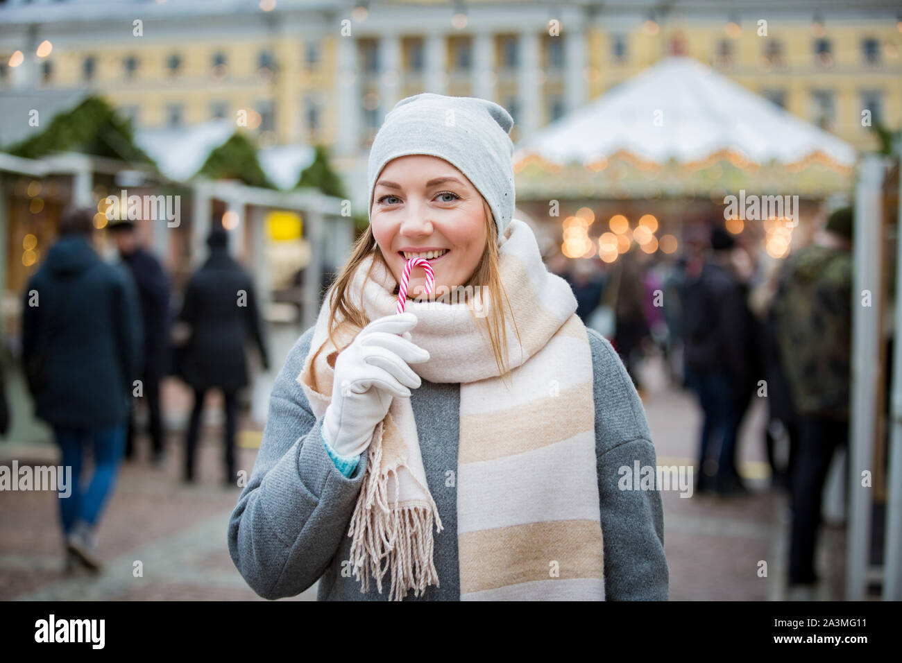 Giovane donna nel mercato di Natale di mangiare candy cane indossare caldo a maglia cappello e sciarpa. Illuminata e decorata fair chioschi, negozi e carosello Foto Stock