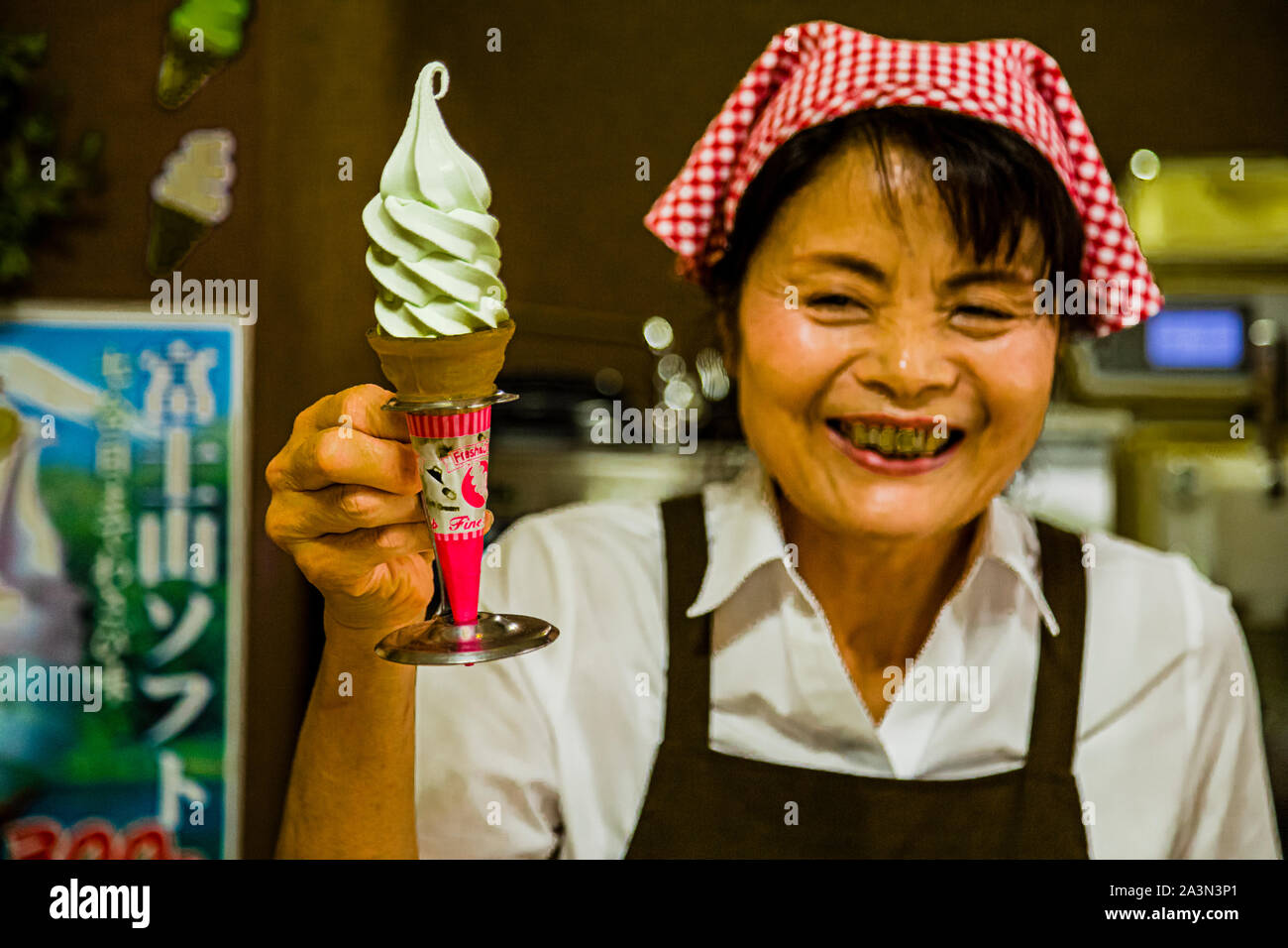 Un'assistente di negozio che ride nel Museo Wasabi di Izu a Kannami, in Giappone, tiene in mano un cono di gelato molle al wasabi verde pallido, mentre un poster in sfondo pubblicizza "Fujisan Soft" (富士山ソフト), gelato molle marchiato dopo il Monte Fuji. Foto Stock