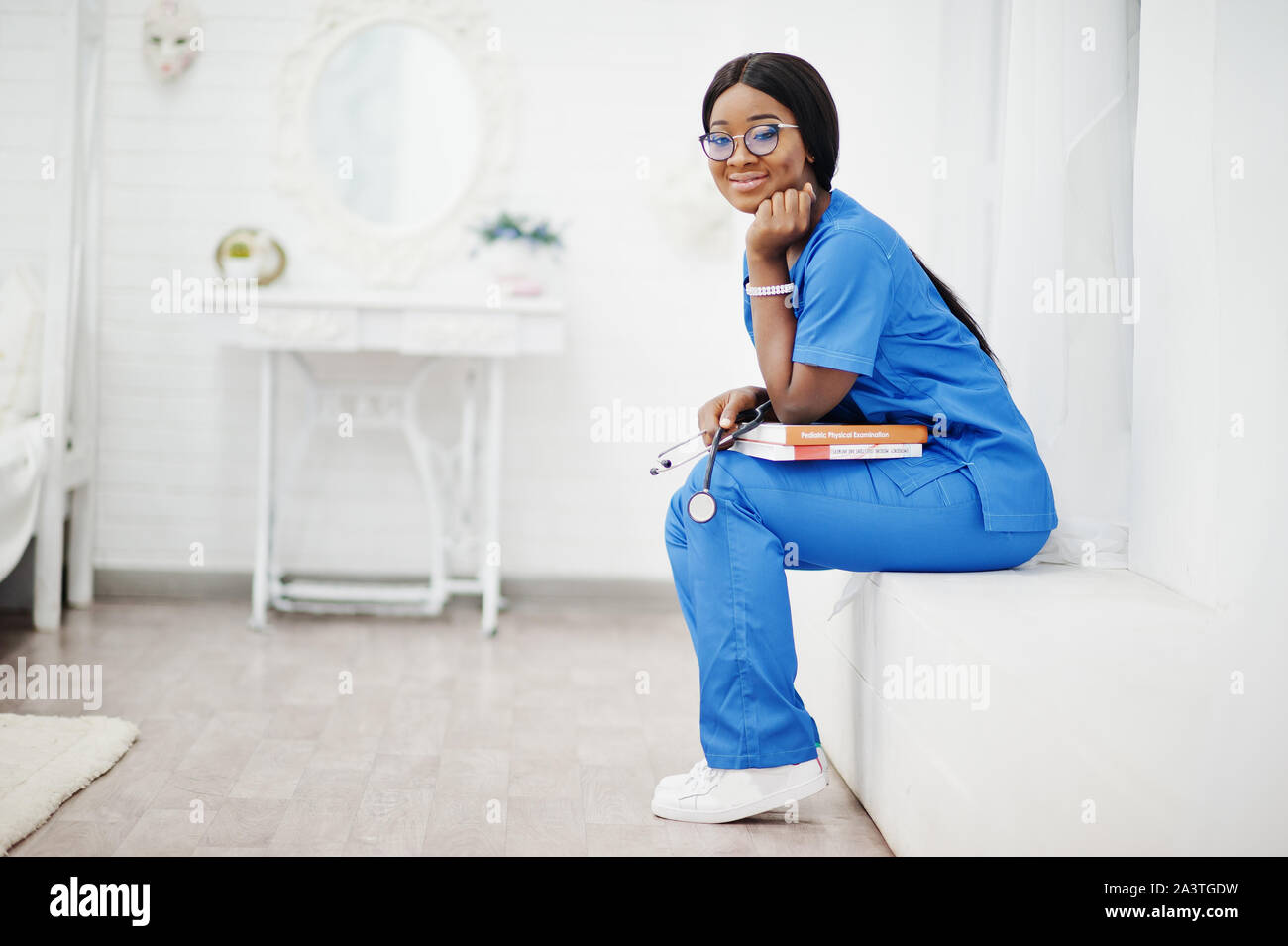 Ritratto di donna felice americano africano giovane medico pediatra in uniforme blu e il cappotto di uno stetoscopio con libri a mani. Sanitario, medico, med Foto Stock