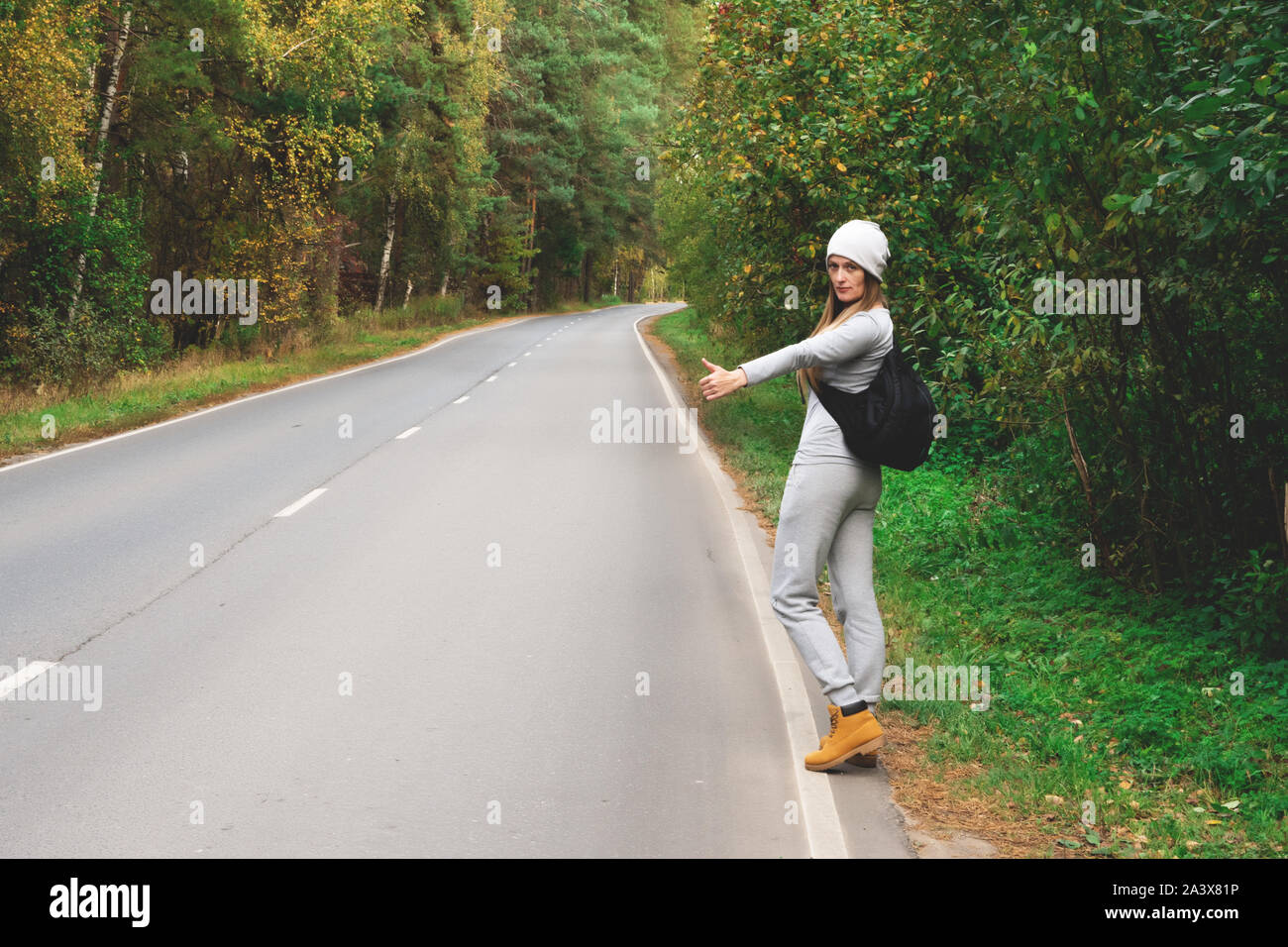 Una giovane e bella ragazza voti sulla strada. Strada d'autunno. La cattura di un auto su una strada vuota. Autostop. Viaggio gratuito in auto Foto Stock