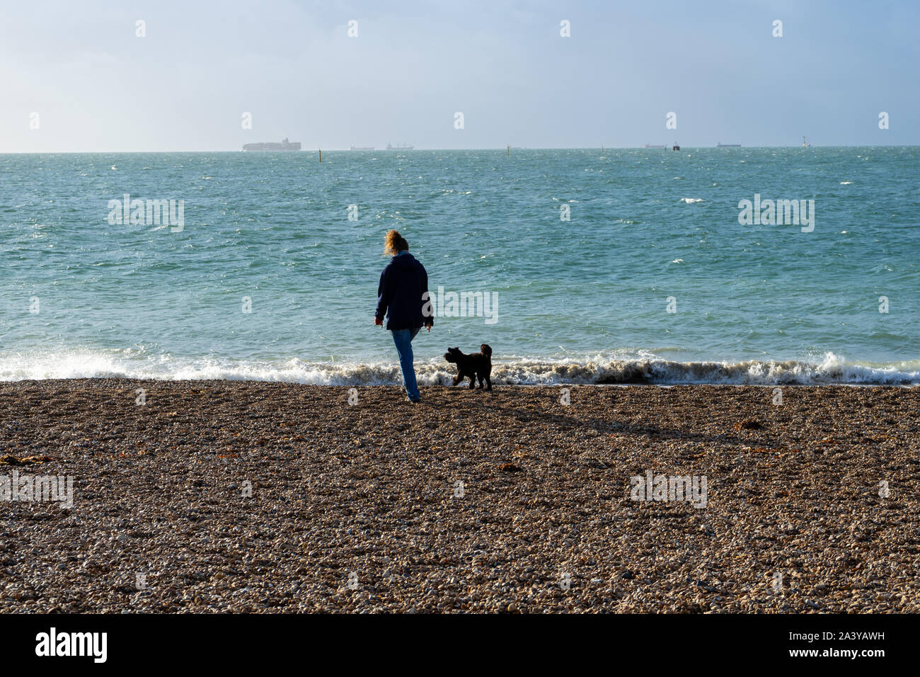 Una donna di mezza età a piedi il suo cane nero lungo una spiaggia di ciottoli con il mare in background Foto Stock