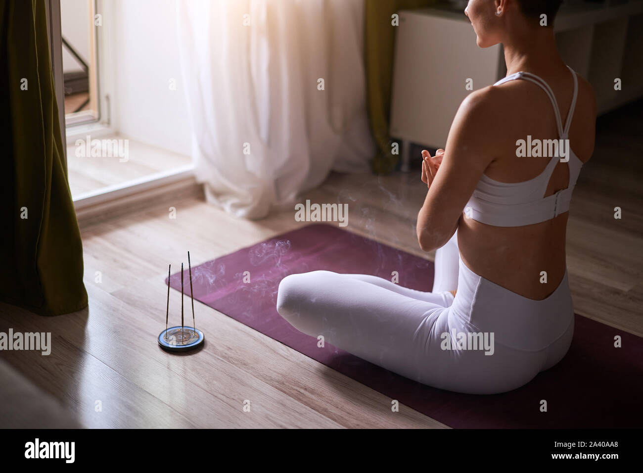 Donna meditando in ambienti chiusi. Attraente e a pelo corto modello caucasici in bianco sportclothes seduta sul tappeto e praticare lo yoga con candele. Pregare Foto Stock