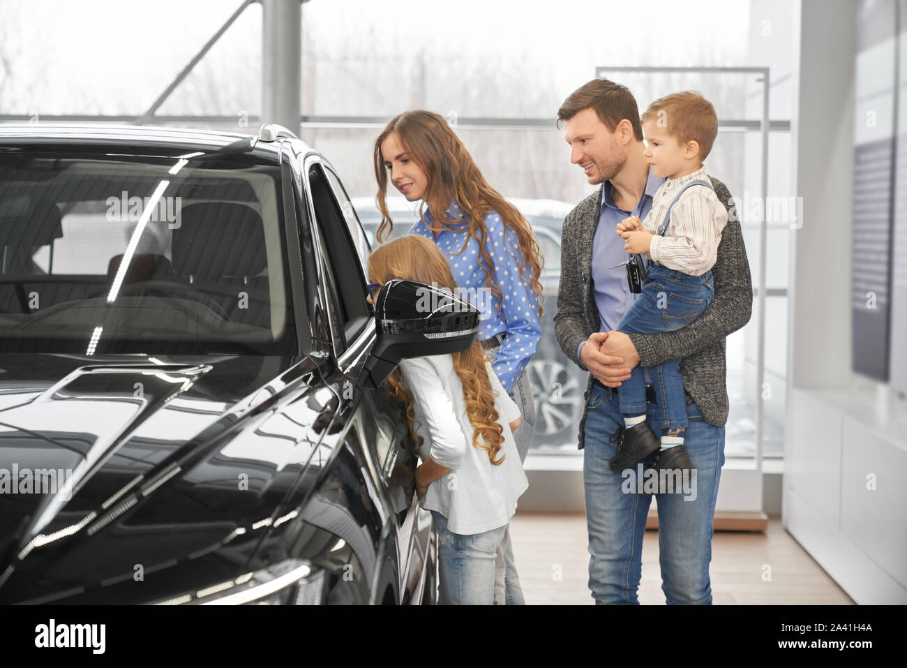 Gentile i bambini con i genitori guardando auto salon attraverso la finestra aperta in showroom. La donna e l uomo con i bambini scegliendo e test vetture alla presentazione. Concetto di acquistare. Foto Stock