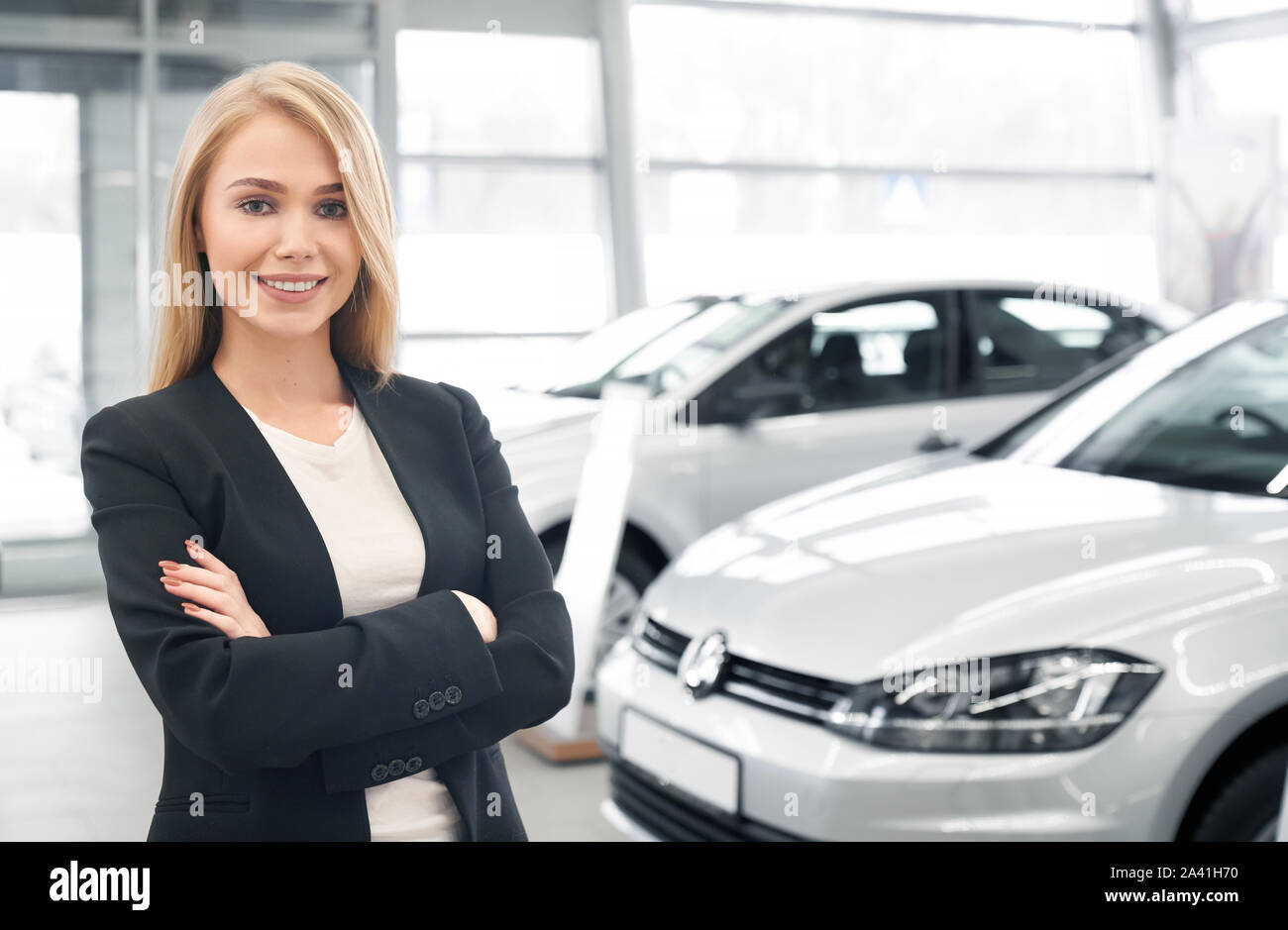 Donna sorridente rivenditore di auto con bracci ripiegati guardando la fotocamera e in posa di fronte a nuove automobili. Bella donna che indossa smart suit lavorando in auto salon e vendita di automobili per i client. Foto Stock